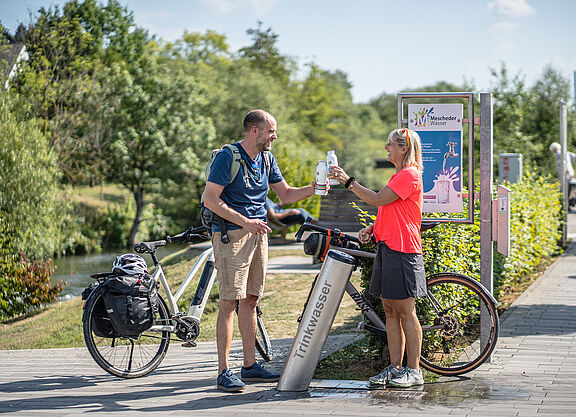 Wasserflaschen auffüllen am RuhrtalRadweg. Zwei Radfahrende füllen Trinkflaschen an einem Trinkbrunnen des RuhrtalRadwegs auf.