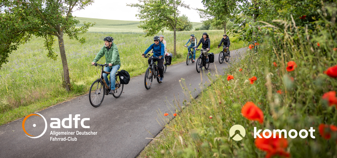 Gruppe Radfahrender auf Radweg zwischen blühenden Wiesen mit Logo ADFC und komoot